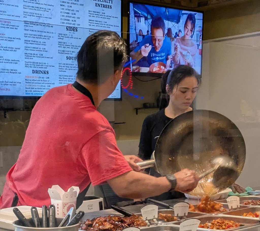 A cook serving orange chicken while looking at a TV. A cashier/server looks down, in her own thoughts